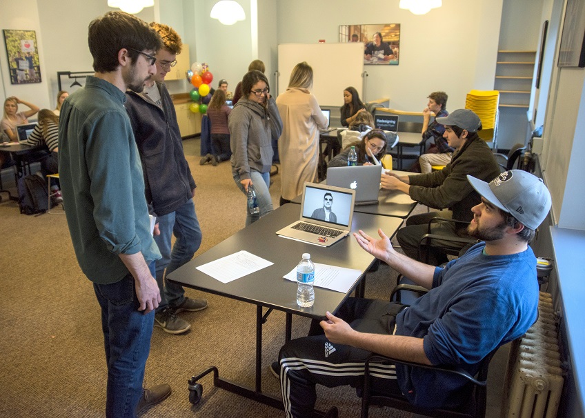 Two students stand before a folding table, a seated student gestures to them with papers and a laptop between them.